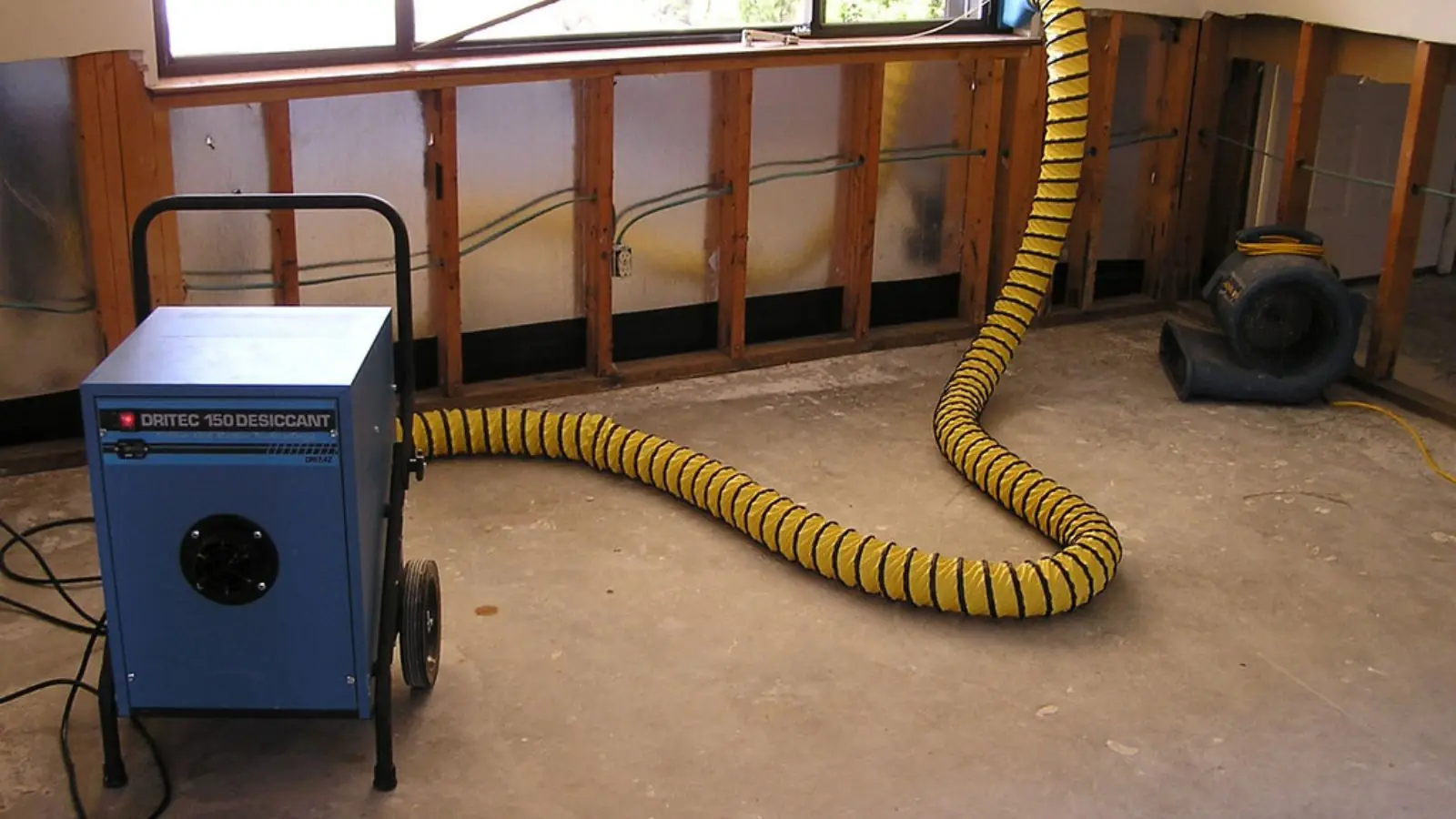 Dehumidifier with a yellow duct drying a room where the lower wall has been opened after water damage